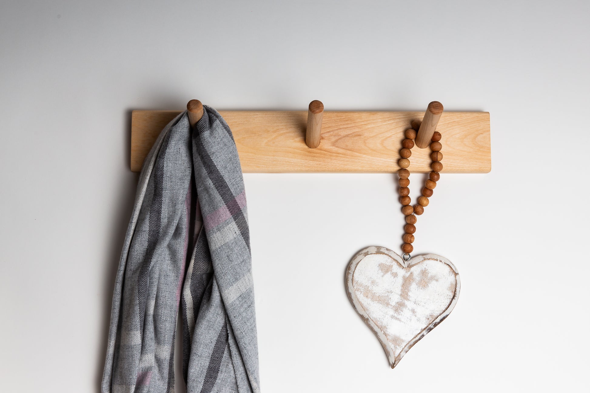 A wooden dowel coat rack with three hooks mounted on a wall, with a coat hanging on one of the hooks and a decorative heart-shaped item hanging on another.