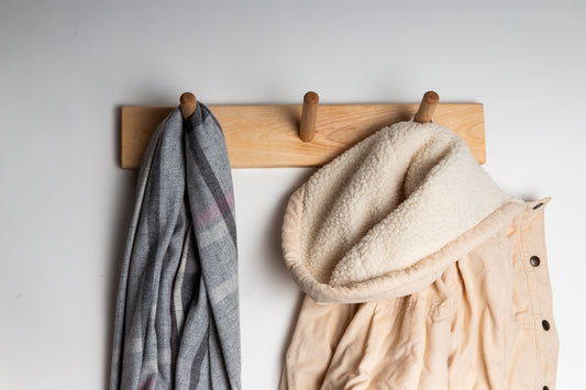 A 3 dowel birch wood wall mounted coat rack with a scarf hanging on the left peg and a corduroy coat hanging on the right peg.