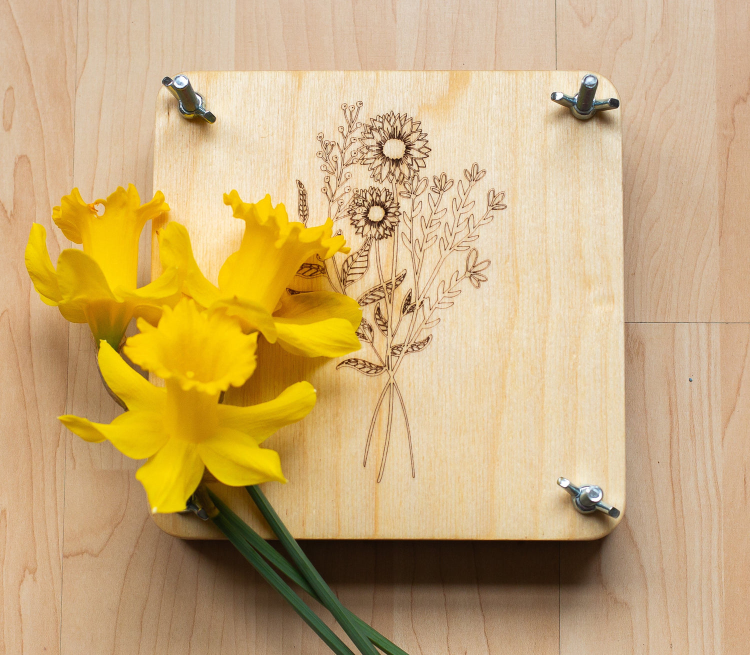 A wooden flower press with a floral design, accompanied by yellow flowers, on a wooden surface.